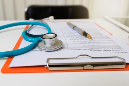 A clipboard with a medical form is displayed on a white desk with a stethoscope and pen.